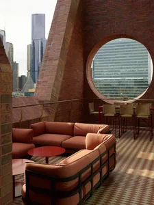 A rooftop lounge area with brick walls, a red circular table, and brown sofas and chairs.