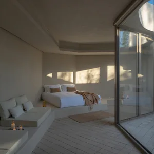 A minimalist bedroom featuring a concrete and glass platform bed, with white bedding and a window allowing sunlight to stream