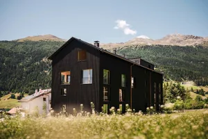 A black wooden building with large windows in a picturesque mountainous landscape.