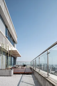 Modern minimalist rooftop patio with wooden deck, glass railings, and potted plants overlooking a cityscape.
