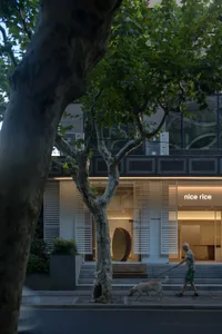 一位行人牽著狗走在人行道上，旁邊有樹木、現代店面與綠意。