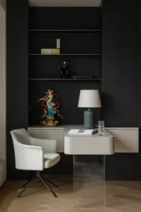 A modern minimalist home office featuring a glass desk, upholstered chair, and bookshelf filled with books and a statue.