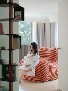 A person holding a white cat sits on a red and white striped sofa in a modern living room with a bookshelf and a window.