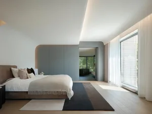 A minimalist bedroom with a bed, pillows, and a rug on a wooden floor, featuring curved architectural elements and large wind