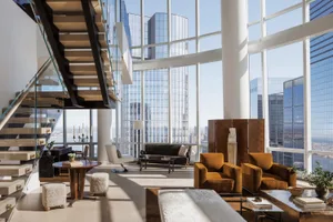 A modern living room featuring large glass windows with a view of the cityscape, wooden furniture, and stylish decor.