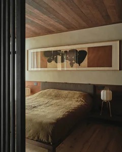 A minimalist bedroom featuring a wooden bed frame and headboard, with a large framed abstract painting above it.