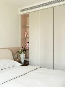 A minimalist bedroom features a bed with white sheets, a beige upholstered headboard, and a white vase with flowers on a nigh