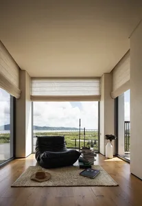 A minimalist living room with a black lounge chair, a stack of books, and a vase with feathers on a wooden floor, featuring l