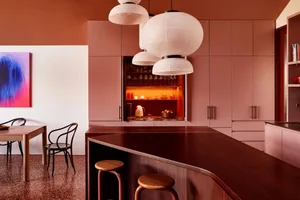 Modern kitchen interior with wooden counter featuring a pink wall and white ceiling, pendant lights hanging above a bar count