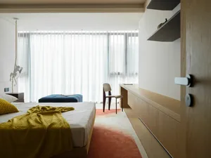 A minimalist bedroom with a wooden floor, bed, and desk, featuring floor-to-ceiling windows with white curtains, a yellow bla