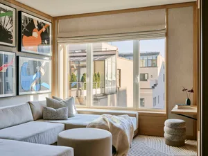 A modern residential interior featuring a fabric window seat with cushions and a blanket, sitting area, and framed artwork on