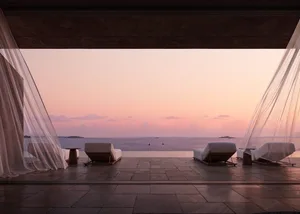 A minimalist outdoor lounge featuring white fabric seating and wooden frames, overlooking the ocean at sunset.
