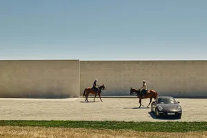 Two people riding horses in front of a concrete wall, with a car parked nearby.
