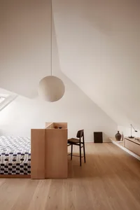A minimalist bedroom featuring a wooden bed, a hanging light fixture, and a chair.