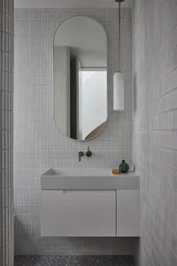 Modern minimalist bathroom featuring grey textured tiles, a sleek white sink with a single faucet, and a large oval mirror ab