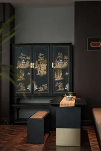 A black lacquer cabinet with gold inlay depicting Chinese landscapes, placed in a darkly painted room with a low table in fro
