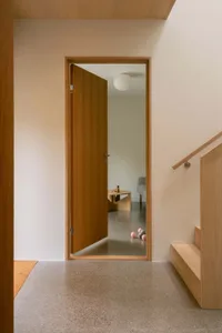 An open wooden door in a minimalist entryway, with a concrete floor, a wooden staircase, and a view into another room with a