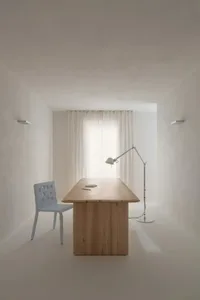 A minimalist office space featuring a wooden desk and chair next to a window with sheer curtains, illuminated by a lamp.