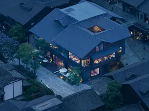 Aerial view of a traditional clay restaurant exterior at dusk, featuring large windows displaying dinner tables, wooden floor