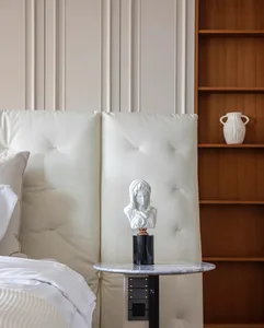A minimalist bedroom featuring a leather headboard with tufted cushions, a marble bedside table, and a wooden bookshelf with 