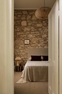 Minimalist bedroom featuring a stone wall, beige carpet, bed with burgundy pillows, wooden stool with lamp, and a woven penda