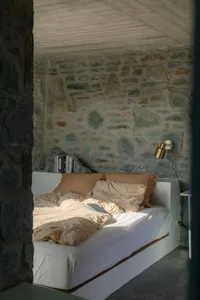 A minimalist bedroom featuring a bed with a stone wall behind it, illuminated by natural light.