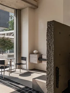 A modern interior with concrete walls and a large window, featuring a black table and chair, and a white vase.