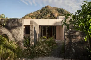 A modernist house with concrete exterior and large windows, situated against a scenic mountain backdrop.