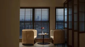 A cozy coffee shop interior featuring wooden cane furniture, a small table, a vase with flowers, and vertical blinds on the w