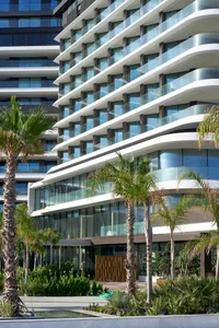 Modern residential building with glass windows and concrete balconies, surrounded by palm trees and greenery.