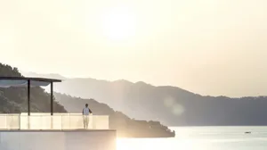 Person standing on a balcony overlooking a serene lake surrounded by mountains during sunset.
