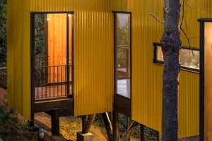 A modern, yellow treehouse with corrugated metal siding and a wooden deck, illuminated from the inside.