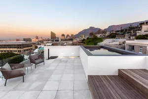 Modern rooftop deck with white concrete tiles, minimal furniture, and a view of the city skyline and mountains at sunset.