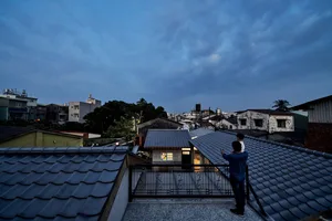 Two people standing on a rooftop with tiled roofs of residential buildings in the background, during an evening setting.
