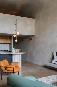 Modern minimalist living room featuring a concrete wall, a turquoise sofa, and orange armchairs, with a kitchen area in the b