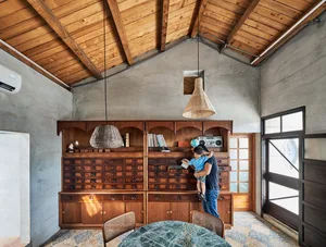 A modern living room featuring wooden furniture, a hanging rattan lamp, and concrete walls.
