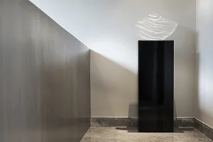 A modern, abstract glass sculpture with a wave-like design displayed on a black pedestal in a minimalist interior setting.