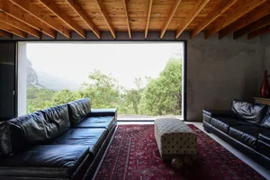 Modern living room with black leather sofas, wooden beams on ceiling, and large window overlooking greenery.