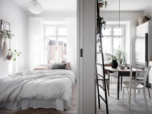 A minimalist bedroom featuring a cozy bed covered in light-colored cotton textiles, surrounded by natural light and green pla