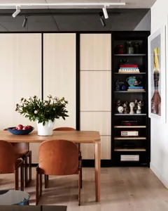 Contemporary dining room featuring a wooden table, glass vase with flowers, and a bookshelf displaying decorative items.