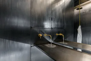 A bathroom with metallic walls and fixtures, featuring stainless steel countertops, gold faucets, and a shower head mounted o