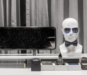 A white mannequin bust with sunglasses and a bow tie, displayed on a white shelf with a black speckled mirror and boxes.