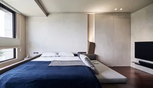 A minimalist bedroom featuring a blue bed with white sheets and pillows, set against minimalist white walls and a wooden floo