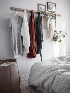 A minimalist bedroom design featuring a bed with white sheets, a wooden coat rack with various clothing items, and a hanging