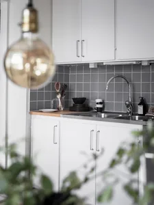 A modern, minimalist kitchen with white cabinets, a stainless steel sink, gray tiled backsplash, and a hanging light bulb.