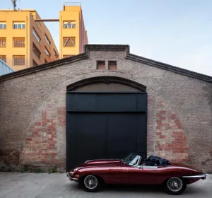 Red classic sports car parked outside a brick building with an arched roof and black gates.