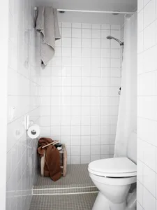 A minimalist bathroom with white tiled walls and floor, a white toilet, and a shower area with a white curtain.