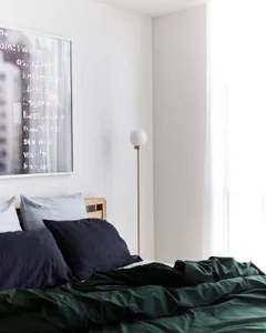 A minimalist bedroom with white walls, white flooring, and white blinds, featuring a bed with dark blue and green bedding, wh