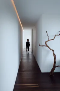 A minimalist interior with white walls and a dark wooden floor, featuring a person walking down a hallway.