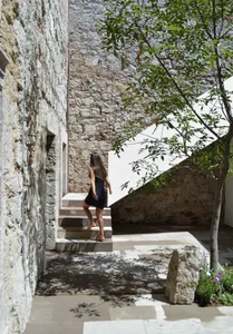 A person in a black dress walks up stone steps surrounded by tree shadows in a courtyard made of stone.